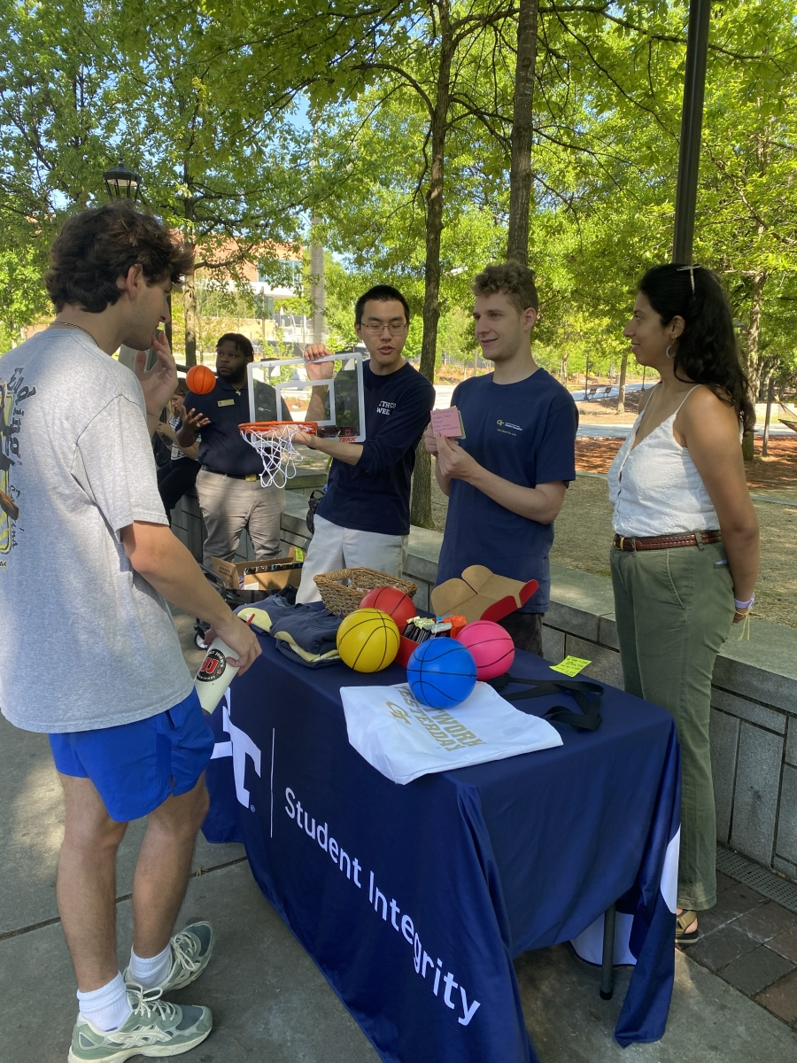 four college students outside under shady trees, standing at a table with toy basketballs