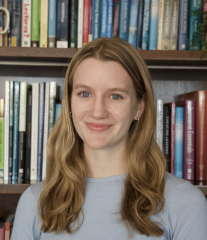 A young woman smiles at the camera. She is seen from the chest up in front of a bookshelf, her wavy blond hair falling over her grey sweater.